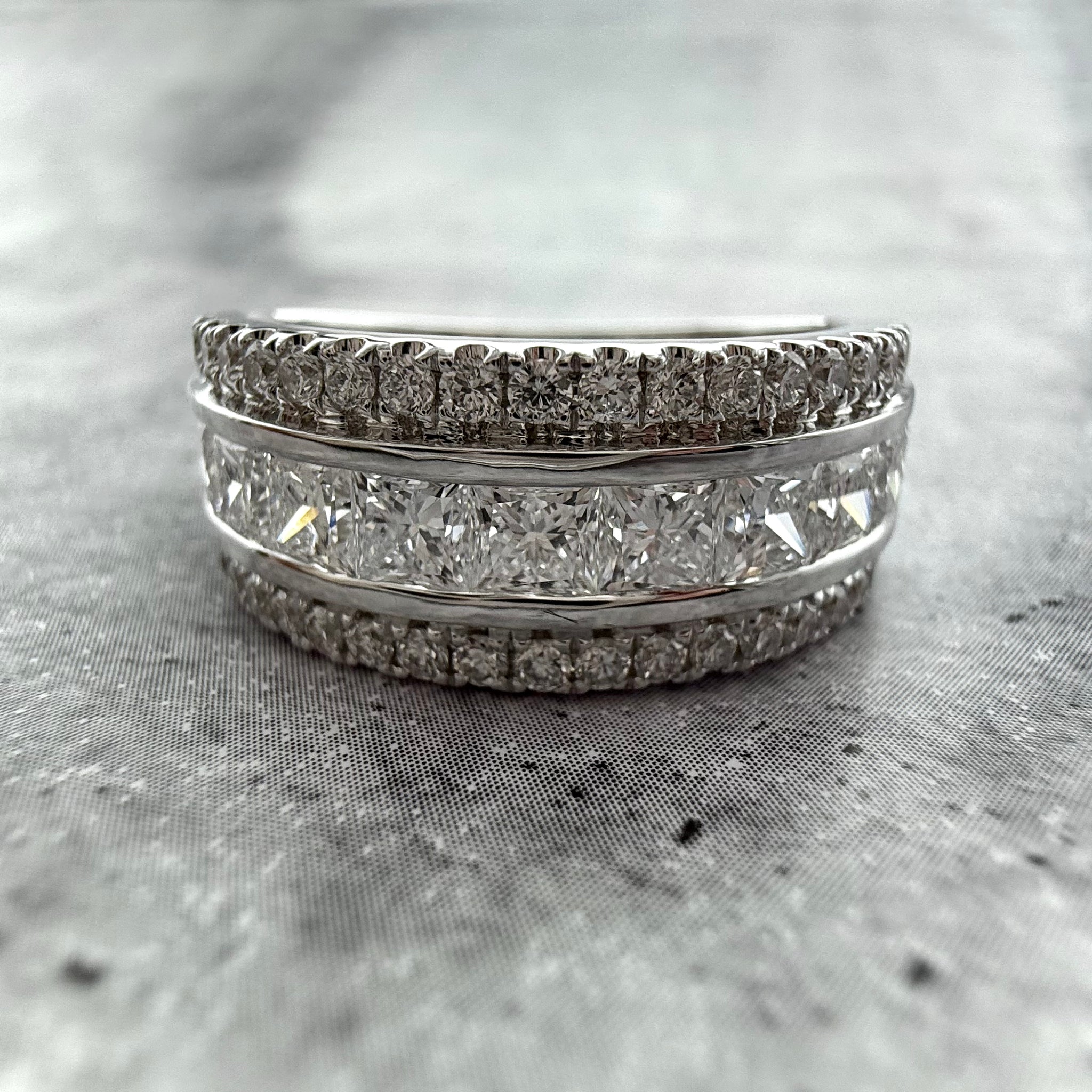 A close up shot of a Custom Wedding band in 14k White gold with princess & round cut diamonds, with a grey textured background