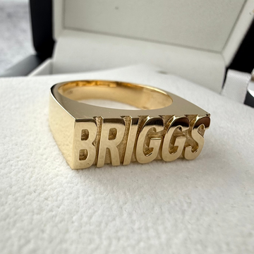 Custom Gold name ring with the writing 'BRIGGS' on the front of the ring laid on a white textured surface