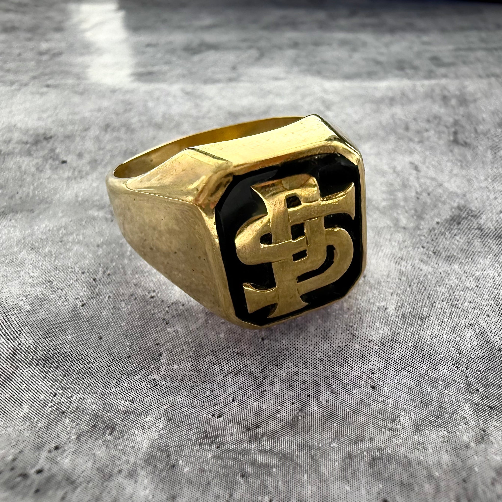 This is a custom initial P & S signet ring in yellow gold with a black enamel background. on a grey textured background