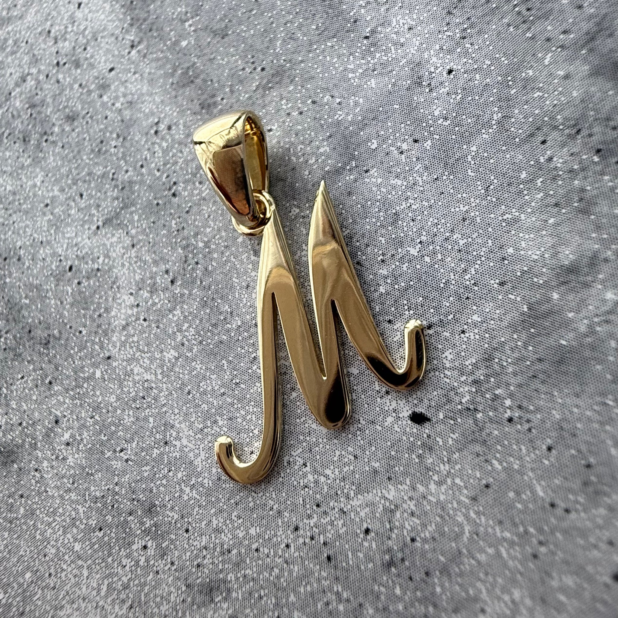 Custom Gold 'M'  initial pendant on a grey textured background