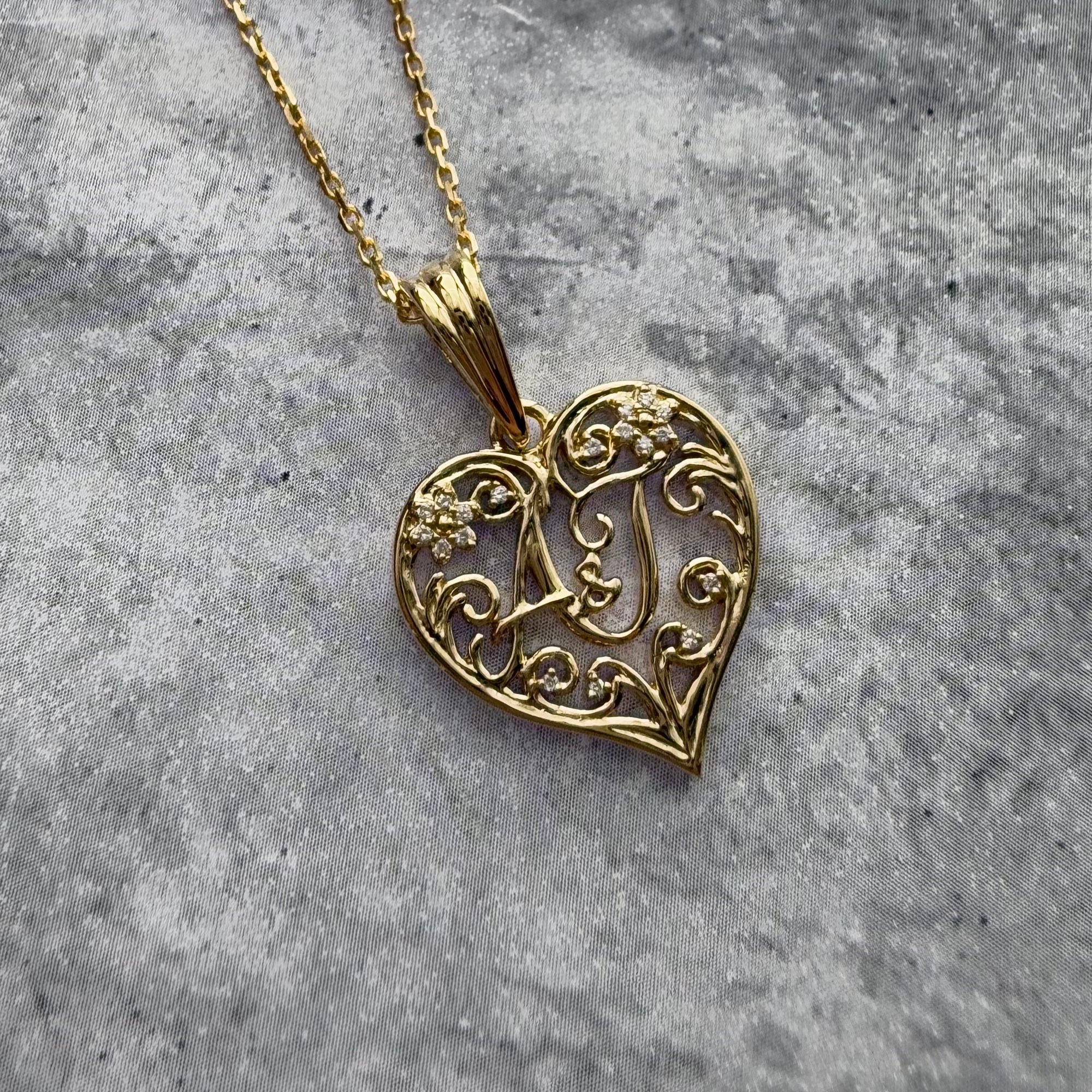 Custom 18k yellow gold A & J Inital heart pendant with a yellow gold cable chain laid on a textured grey background.