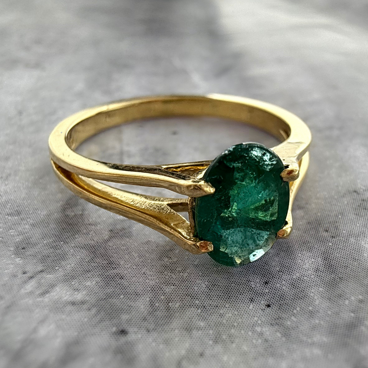 Side Close up shot of a Custom 18k Yellow gold 4prong Emerald gemstone ring, laid on a grey textured background.