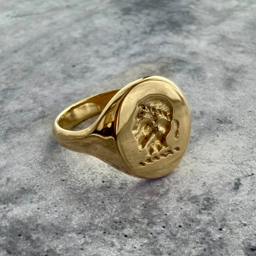 A close up of a crest signet ring in yellow gold. the background is a grey textured background