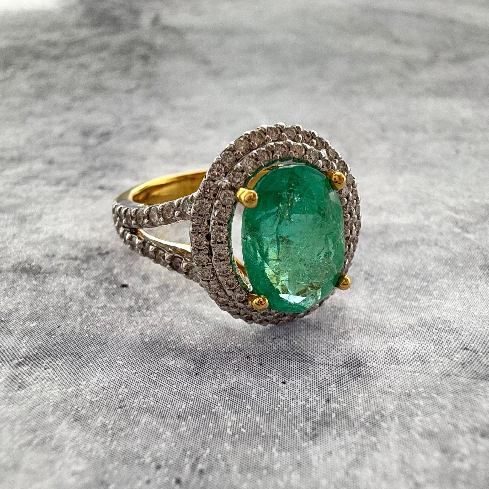 A close up shot of a custom 18k yellow gold oval Cut emerald ring, with a double halo of diamonds. with the background being textured and grey in colour.