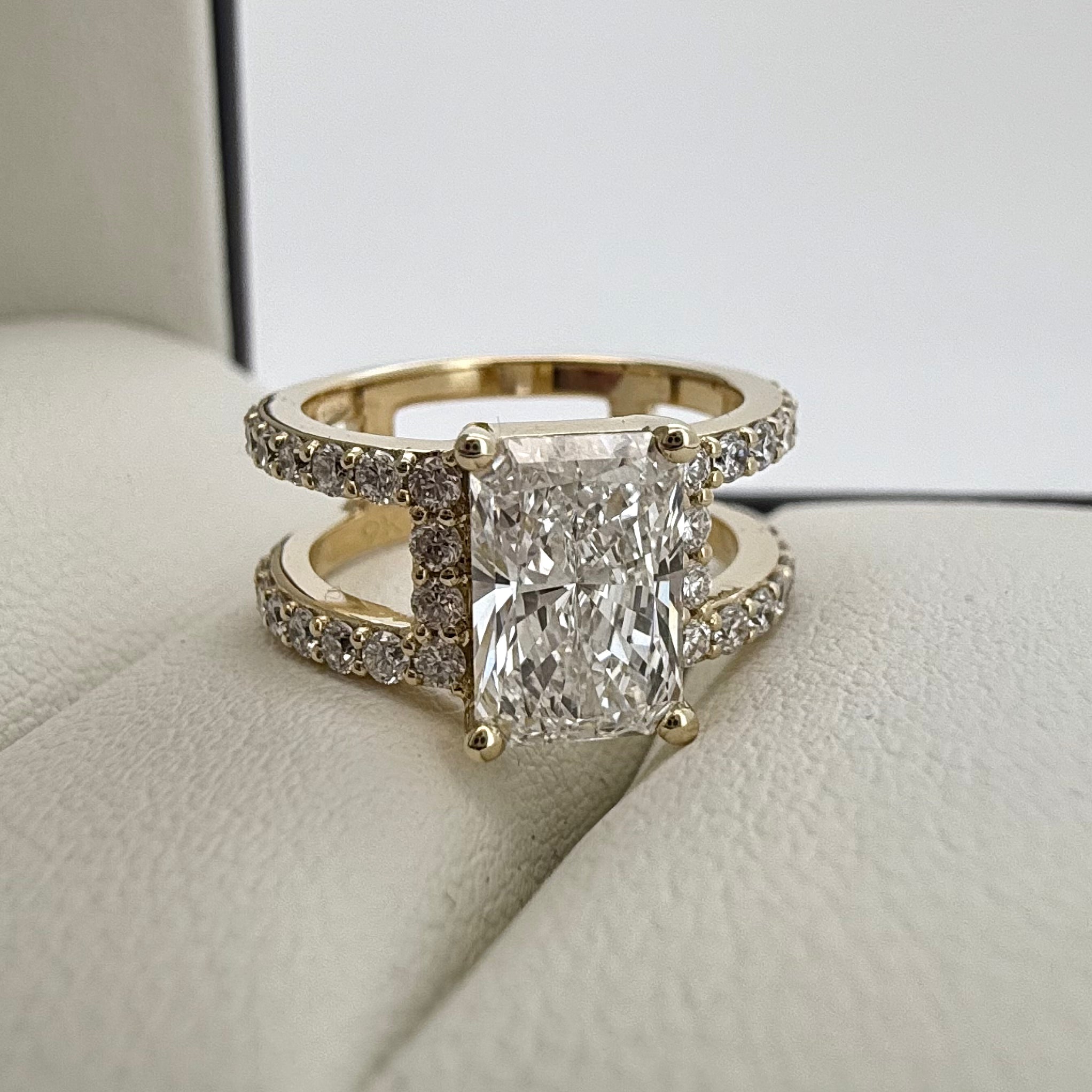 Close up front shot of a custom Yellow gold Radiant cut Diamond Engagement ring, with side diamonds, laid on top of a cream textured cushion